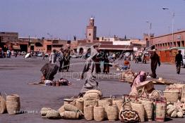 Image du Maroc Professionnelle de  Une femme étale ses paniers en osier à même le sol sur la fameuse place Jemaa El Fana de Marrakech, en espérant le passage de touristes, mais à cette heure où le soleil est au zénith le lieu est presque vide, s'abriter du soleil est une nécessité pour un bon nombre de résidents de la ville touristique du Maroc, au fond le minaret de la Mosquée Kharbouche, Jeudi 19 Mai 1988. (Photo / Abdeljalil Bounhar) 